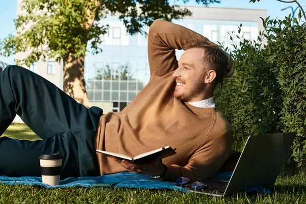 Genç bir adam açık havada bir battaniyede dinleniyor, kitap okuyor ve yakınlarda bir kahve fincanıyla gülümsüyor..