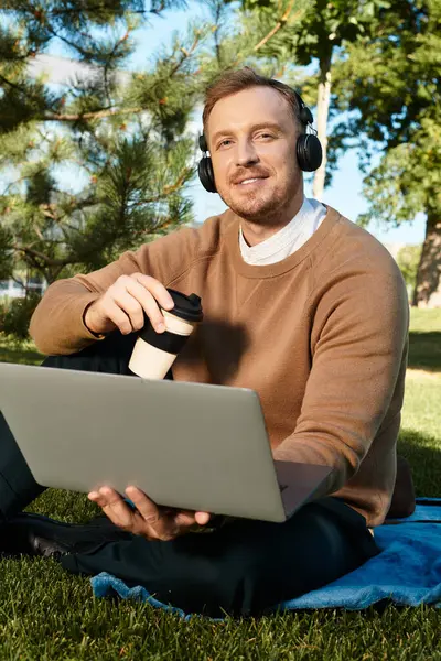 Genç bir adam, bir üniversite öğretmeni, çimlere dizüstü bilgisayar ve kahve fincanıyla oturmuş, mutlu bir şekilde gülümsüyor..