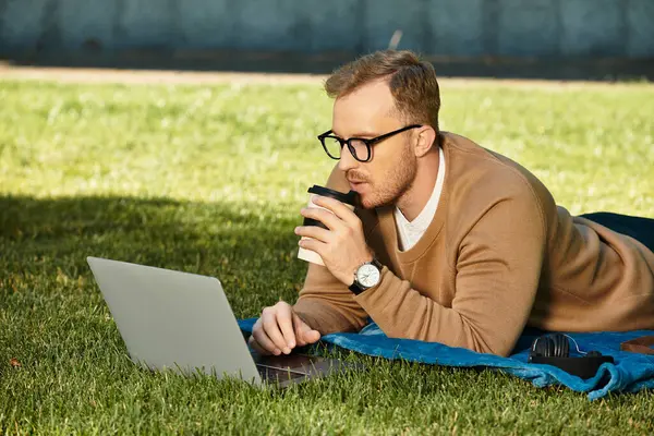 Genç bir öğretmen, dizüstü bilgisayarını kullanarak çimlerin üzerinde rahatlar ve güneşin altında kahve yudumlar..