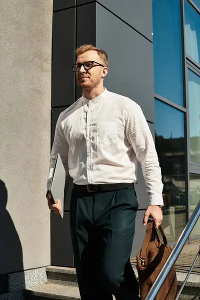 Akıllı giyimli genç bir adam, modern bir binadan elinde laptop ve çantayla ayrılır..