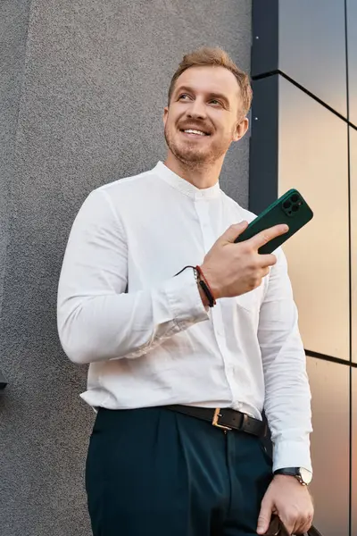 Şık bir gömlek ve pantolon giymiş genç bir adam dışarıda duruyor, telefonunu tutuyor ve gülümsüyor..