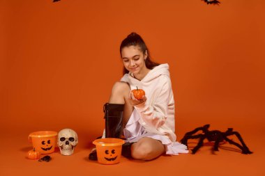 Neşeli bir kız, canlı turuncu bir arka planda küçük bir balkabağıyla oynayarak Cadılar Bayramı 'nı kutluyor..