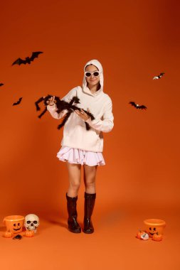 Tatlı bir kız, turuncu bir arka planda Cadılar Bayramı eğlencesini sever. Etrafı ürkütücü dekorasyonlarla çevrili..