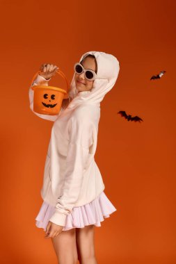 Samimi kıyafetli bir kız, Cadılar Bayramı 'nı neşeli bir kovayla turuncu bir arka planda kutluyor..