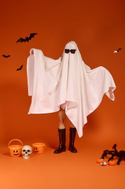 Bir çocuk neşeli bir şekilde hayalet kostümü ve güneş gözlüğünü turuncu bir arka fona takar..
