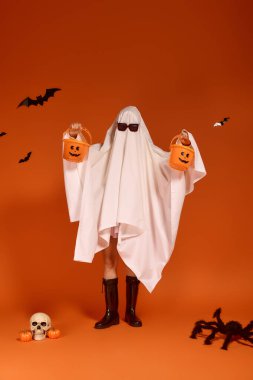 Hayalet kostümü giyen oyuncu bir kız turuncu bir arka fonun önünde balkabağı kovaları tutuyor..