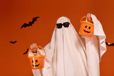 Hayalet kostümü giymiş neşeli bir çocuk elinde turuncu şeker kovalarıyla Cadılar Bayramı 'nı neşeyle kutluyor..