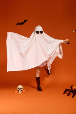 Hayalet kostümlü bir çocuk, parlak turuncu bir arka planda Cadılar Bayramı 'nı kucaklayarak mutlu bir şekilde dans eder..