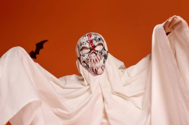 Hayalet gibi giyinmiş neşeli bir kız canlı turuncu bir arka planda Cadılar Bayramı ruhunu kucaklıyor..