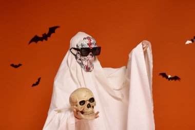 Korkunç bir cadılar bayramı kıyafeti giymiş bir çocuğun elinde turuncu bir arka planda yarasalarla çevrili bir kafatası var..