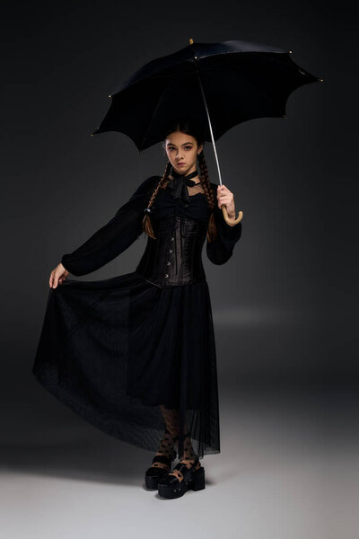 A girl in a gothic Wednesday costume stands confidently, holding a black umbrella.