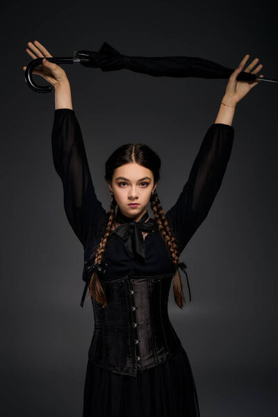 A girl with braids wears a Wednesday dress and holding black umbrella, embodying Halloween vibes.