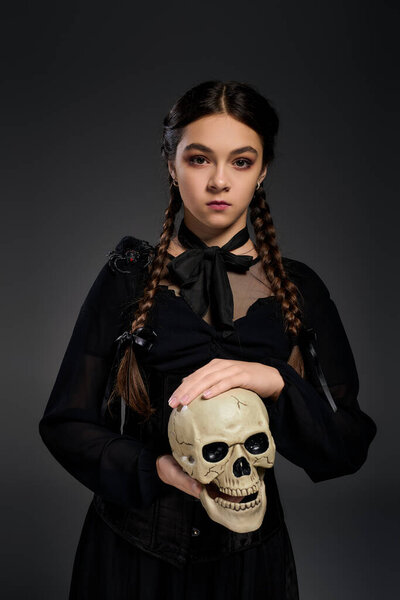 A girl with braids dressed in a gothic style Wednesday dress poses with a skull for Halloween.