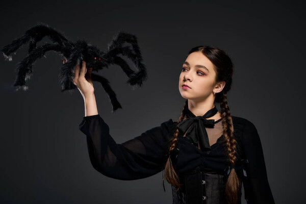 A girl with braids wears a gothic Wednesday costume, holding a spooky spider in a dark setting.
