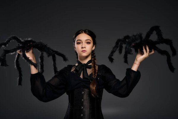 A young girl with braids wears a Wednesday dress and holds spooky spiders, celebrating Halloween.