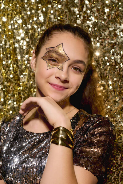 A girl with a star drawn on her cheek poses cheerfully among shiny golden sequins during Halloween.