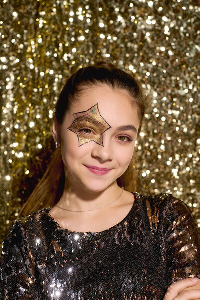 Молодая девушка показывает креативный дизайн звезды на своем лице в окружении мерцающих золотых блёсток.
