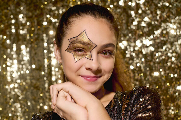 A young girl smiles while showcasing a star on her cheek, sparkling golden sequins around her.