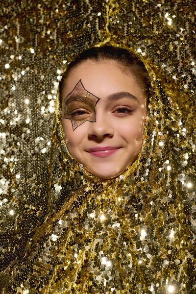 Радостная девушка со звездой, нарисованной на ее лице, ярко улыбается, окруженная золотыми блестками.