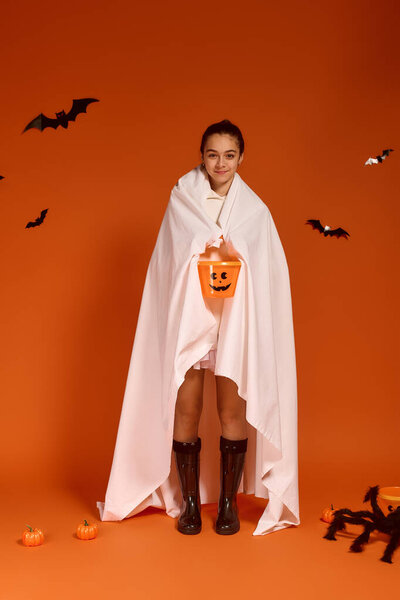 A joyful girl in a white blanket holds a pumpkin bucket, celebrating Halloween in autumn.