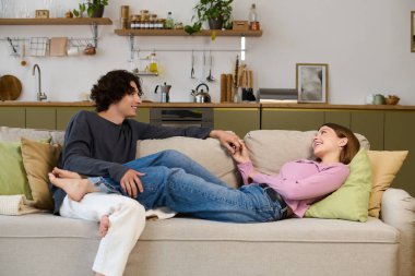 A curly-haired man and a beautiful woman laugh joyfully on their couch in a chic space.