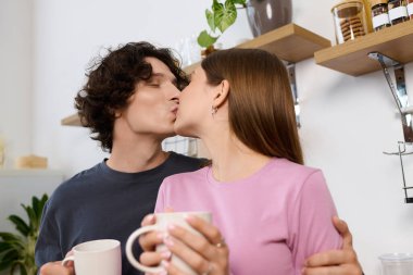 Joyful young couple kisses while holding mugs in their stylish apartment filled with plants.