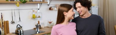 A young couple smiles at each other, enjoying their time in a modern kitchen space, banner