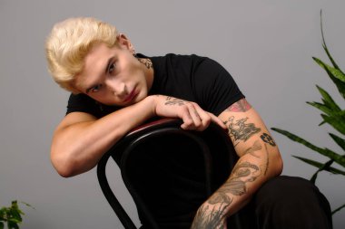 Şık, sarı saçlı genç bir adam sandalyede rahatlıyor, modern gençlik kültürünü somutlaştırıyor..