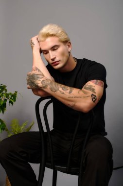 Dövmeleri olan yakışıklı bir genç, minimalist bir stüdyoda sandalyede düşünceli bir şekilde poz veriyor..