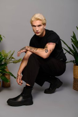 Süslü bitkilerin yanında çömelmiş, sarı saçlı, modaya uygun bir genç..