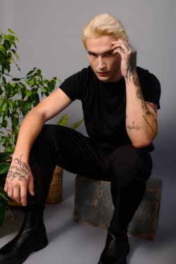 Kendine güvenen genç adam zarif pozlar veriyor. Etrafı canlı yeşilliklerle çevrili şık dövmeleri var..