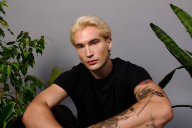 Göz alıcı özellikleri olan şık bir genç, yemyeşil bitkilerin arasında oturur, büyüleyici ve kendine güvenlidir..