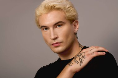 Sarışın ve dövmeli yakışıklı genç, kendinden emin bir şekilde poz veriyor, gençlik kültürünü yansıtıyor..