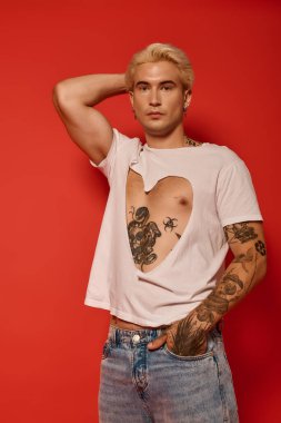 Kalp kesimi gömlekli genç bir adam, bir eli kafasında, dövmelerini renklendiriyor..