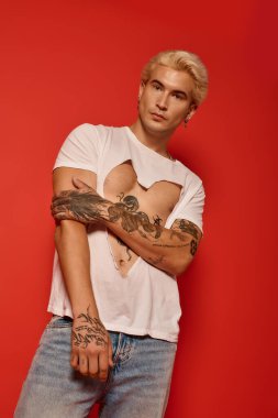 Dövmeleri olan şık bir genç, kırmızı bir arkaplanda kalp kesme gömleğiyle rahatça poz veriyor..