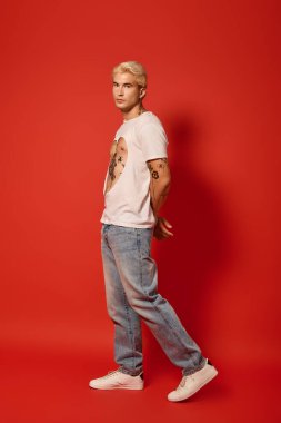 Dövmeli genç cesur bir kırmızı arka plana karşı duruyor, gençlik tarzını sergiliyor..