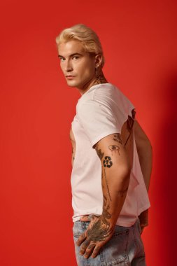 Çarpıcı dövmeleri ve modaya uygun kıyafetleri olan genç bir adam kırmızı arka planda modern gençlik tarzını somutlaştırıyor..