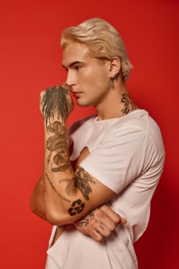 Çarpıcı dövmeleri olan genç yakışıklı bir adam cesur bir kırmızı zemine karşı düşünceli bir şekilde poz verir..