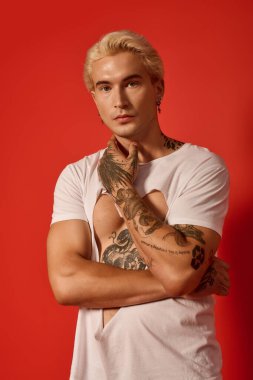 Şık bir genç kollarını kavuşturmuş, çekici bir ifade takınıyor, dövmeleri var..