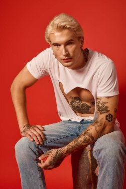 Yakışıklı genç, kendine güvenerek oturuyor, cesur kırmızı bir arka plana karşı şık giyiniyor..