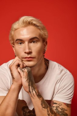 Benzersiz dövmeleri olan çarpıcı sarı saçlı bir genç dikkatle izleyiciye bakıyor..