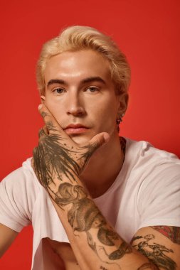 Çarpıcı dövmeleri olan yakışıklı bir genç cesur bir kırmızı duvarın önünde düşünceli bir şekilde poz veriyor..