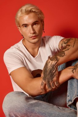 Dövmeleri olan yakışıklı bir genç enerjik kırmızı bir arka plana karşı gevşiyor, gençlik tarzını somutlaştırıyor..