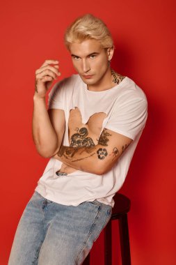 Genç yakışıklı bir adam, bir taburede otururken dövmelerini gösteriyor, modaya uygun bir hava yayıyor..