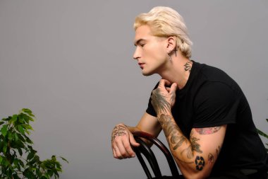 Şık bir genç, sade bir zemine karşı düşünceli bir şekilde poz veriyor, dövmelerini sergiliyor..