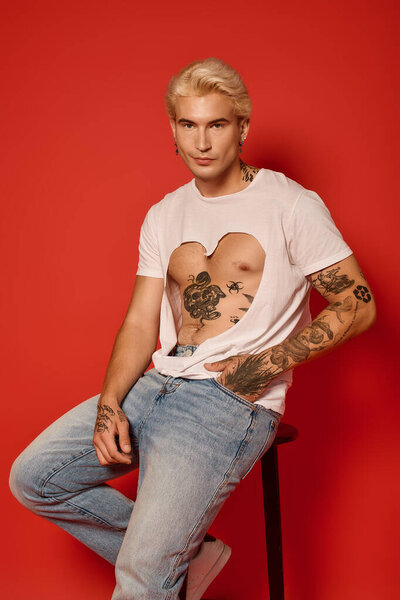 Young man with blonde hair and tattoos poses confidently against a bright red background.