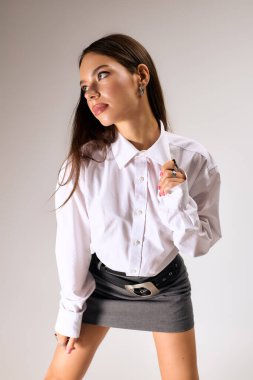 A young woman confidently poses in a trendy autumn outfit in a grey studio.