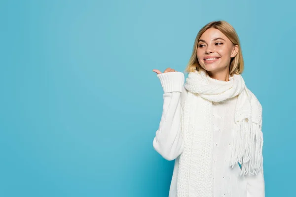 Cheerful blonde woman in white sweater and scarf pointing away with thumb isolated on blue — Stock Photo
