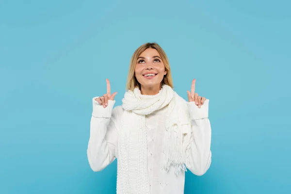 Mujer rubia alegre en suéter blanco y bufanda apuntando hacia arriba con los dedos aislados en azul - foto de stock