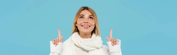 Mulher loira alegre em suéter branco e cachecol apontando para cima com os dedos isolados em azul, banner — Fotografia de Stock